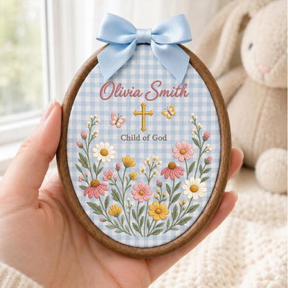 May include: An oval wooden embroidery hoop featuring a light blue and white gingham fabric background. The design includes embroidered pink, yellow, and white wildflowers, a gold cross, two butterflies, and the text Olivia Smith Child of God.