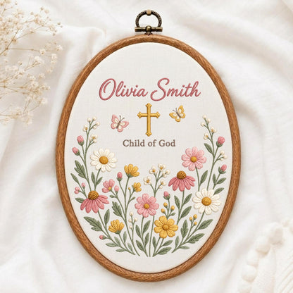 May include: An oval wooden embroidery hoop containing white fabric with the embroidered text Olivia Smith and Child of God. A gold cross sits between two small butterflies, surrounded by a colorful floral arrangement of pink, yellow, and white flowers.