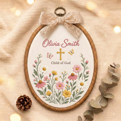 May include: An oval wooden embroidery hoop containing white fabric with a floral design, a gold cross, two butterflies, and the text "Olivia Smith" and "Child of God" embroidered in pink and gray. A beige lace bow is attached to the top.