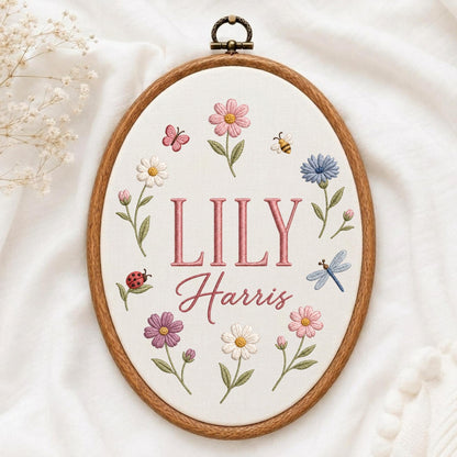 May include: An oval wooden embroidery hoop containing white fabric with the embroidered name LILY Harris in pink. The design features colorful stitched flowers, a ladybug, a butterfly, a bee, and a dragonfly arranged around the text.