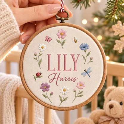 May include: A circular wooden embroidery hoop containing cream-colored fabric with the embroidered name LILY Harris in pink. The design features a floral wreath with pink, white, purple, and blue flowers, a ladybug, a butterfly, a bee, and a dragonfly.