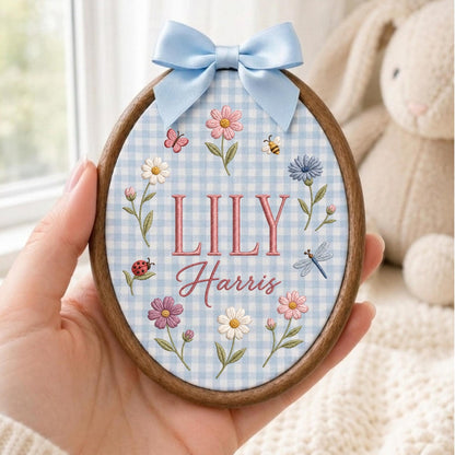 May include: An oval wooden embroidery hoop containing light blue and white gingham fabric. The fabric features embroidered flowers, a ladybug, a butterfly, a bee, and a dragonfly. The text LILY Harris is embroidered in pink in the center.