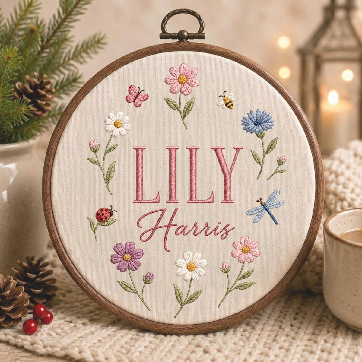 May include: A circular wooden embroidery hoop containing cream-colored fabric with the embroidered name LILY Harris in pink. The design features a floral wreath with pink, white, blue, and purple flowers, a ladybug, a bee, and a dragonfly.