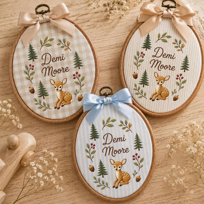 May include: Three oval wooden embroidery hoops featuring fabric backgrounds with a fawn, pine trees, acorns, and foliage. Each hoop displays the name Demi Moore in script and is topped with a satin ribbon bow in beige or light blue.