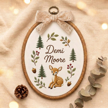May include: An oval wooden embroidery hoop containing white fabric with the embroidered name Demi Moore, a brown fawn, green pine trees, acorns, and leafy branches. A beige lace bow is attached to the top of the hoop with a metal hanger.