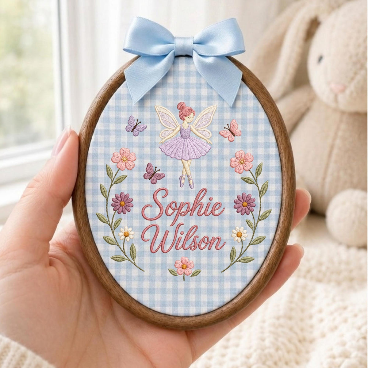 May include: An oval wooden embroidery hoop frame containing light blue and white gingham fabric. The fabric features an embroidered fairy in a purple dress, butterflies, flowers, and the name Sophie Wilson in pink script with a light blue satin bow.
