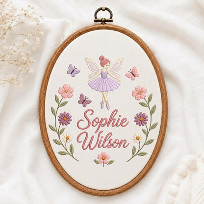 May include: An oval wooden embroidery hoop containing white fabric with a hand-stitched design of a fairy in a purple dress, butterflies, and pink and purple flowers. The text "Sophie Wilson" is embroidered in pink script in the center.