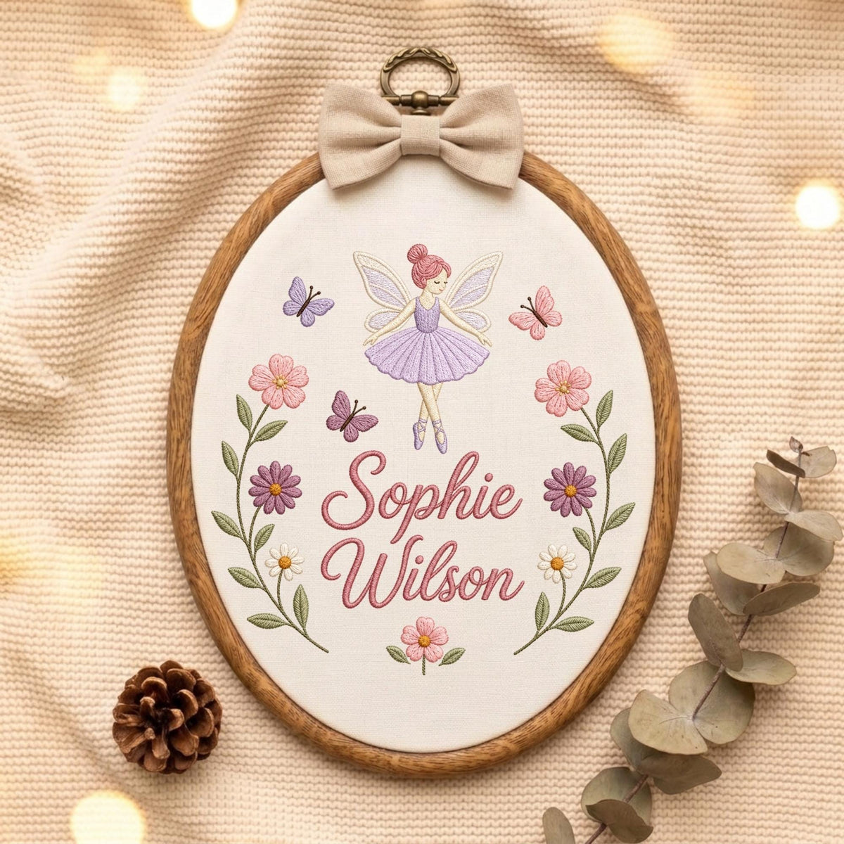 May include: An oval wooden embroidery hoop containing white fabric with a stitched design of a fairy in a purple dress, butterflies, and flowers. The name Sophie Wilson is embroidered in pink script. A beige fabric bow sits at the top.