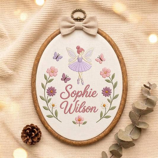 May include: An oval wooden embroidery hoop containing white fabric with a stitched design of a fairy in a purple dress, butterflies, and flowers. The name Sophie Wilson is embroidered in pink script. A beige fabric bow sits at the top.