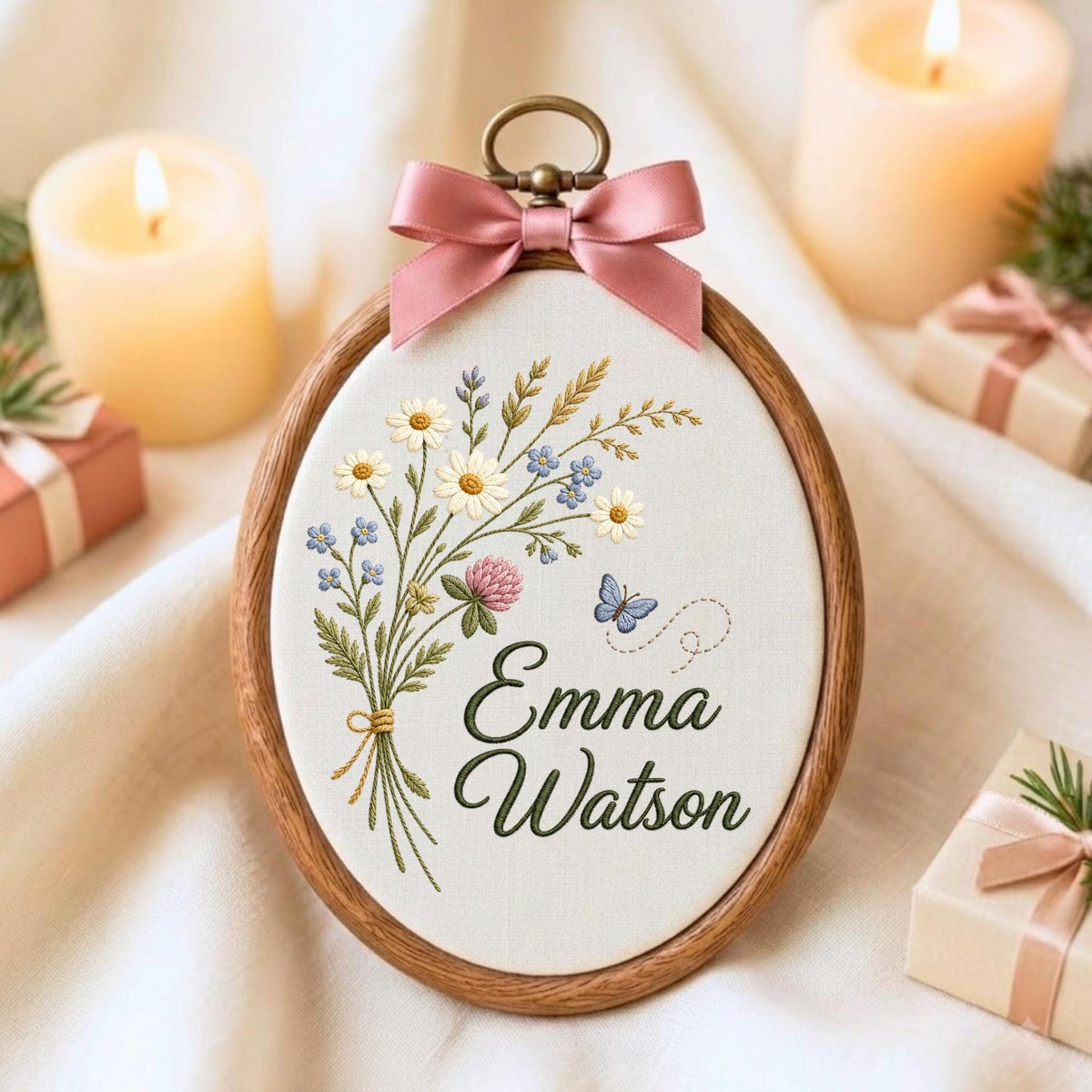 May include: An oval wooden embroidery hoop containing white fabric with a stitched bouquet of daisies, blue flowers, and a clover, accompanied by a small blue butterfly. The name Emma Watson is embroidered in green script below a pink satin bow.