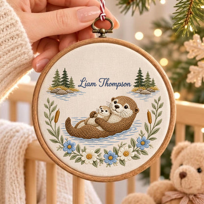 May include: A round wooden embroidery hoop containing a fabric panel with a hand-stitched design of two brown otters floating in water. The text Liam Thompson is embroidered in blue above the otters, framed by green pine trees and flowers.