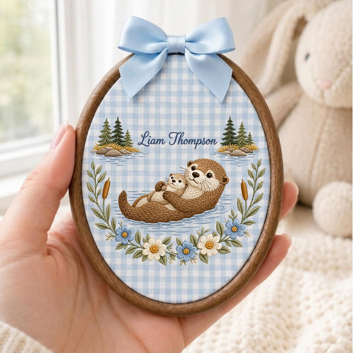 May include: An oval wooden embroidery hoop featuring a light blue and white gingham fabric background. The design shows two brown otters, pine trees, and floral accents. The text Liam Thompson is printed in blue script above the otters.