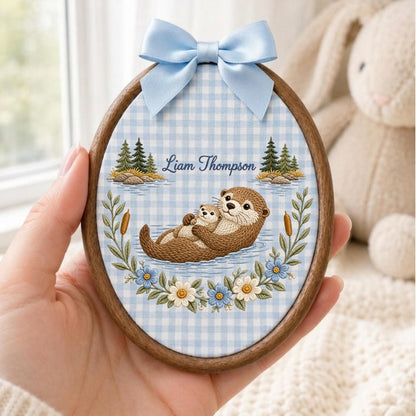 May include: An oval wooden embroidery hoop featuring a light blue and white gingham fabric background. The design shows two brown otters, pine trees, and floral accents. The text Liam Thompson is printed in blue script above the otters.