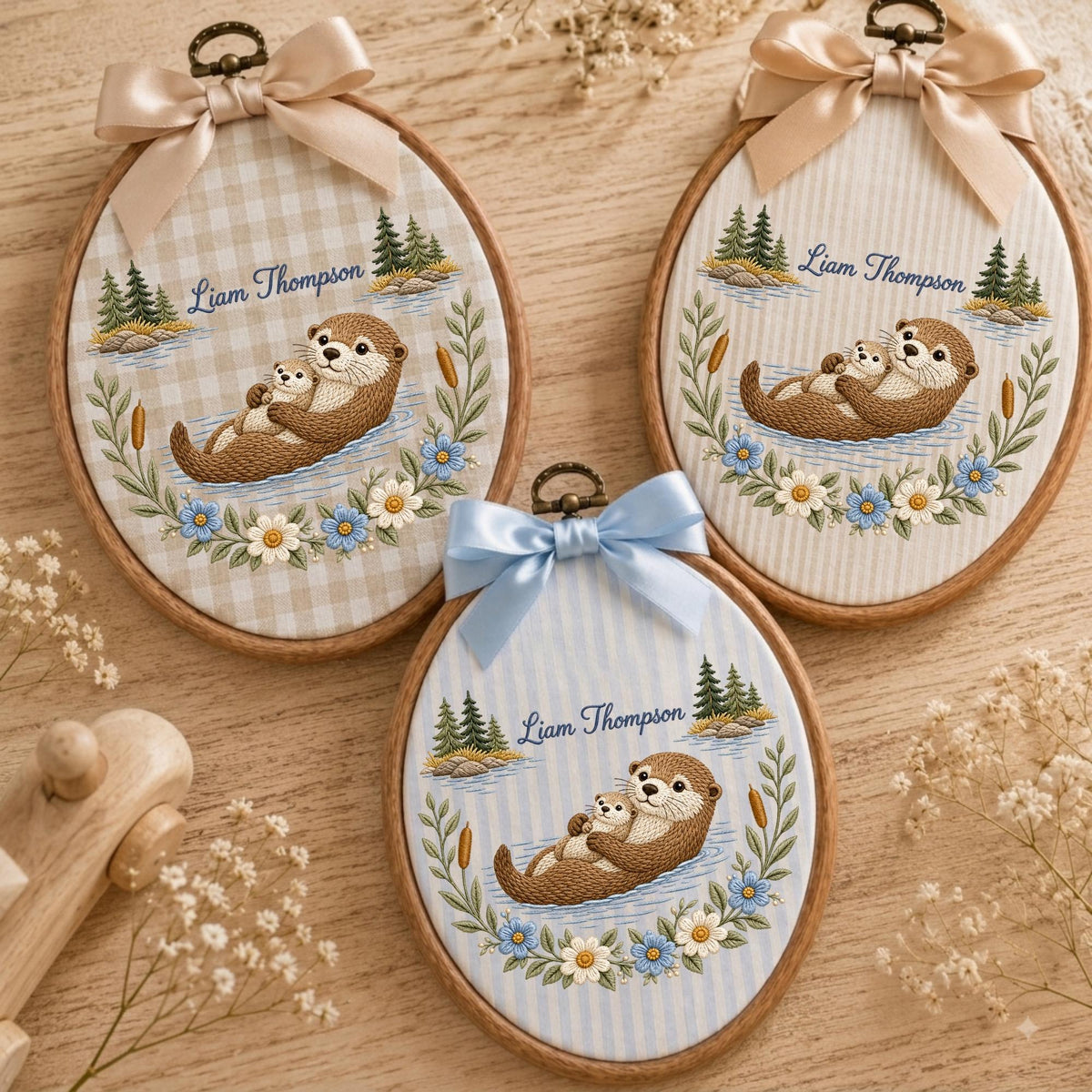May include: Three oval wooden embroidery hoops featuring fabric with a printed illustration of two otters, pine trees, and flowers. Each hoop is topped with a satin ribbon bow and displays the custom name Liam Thompson in cursive script.