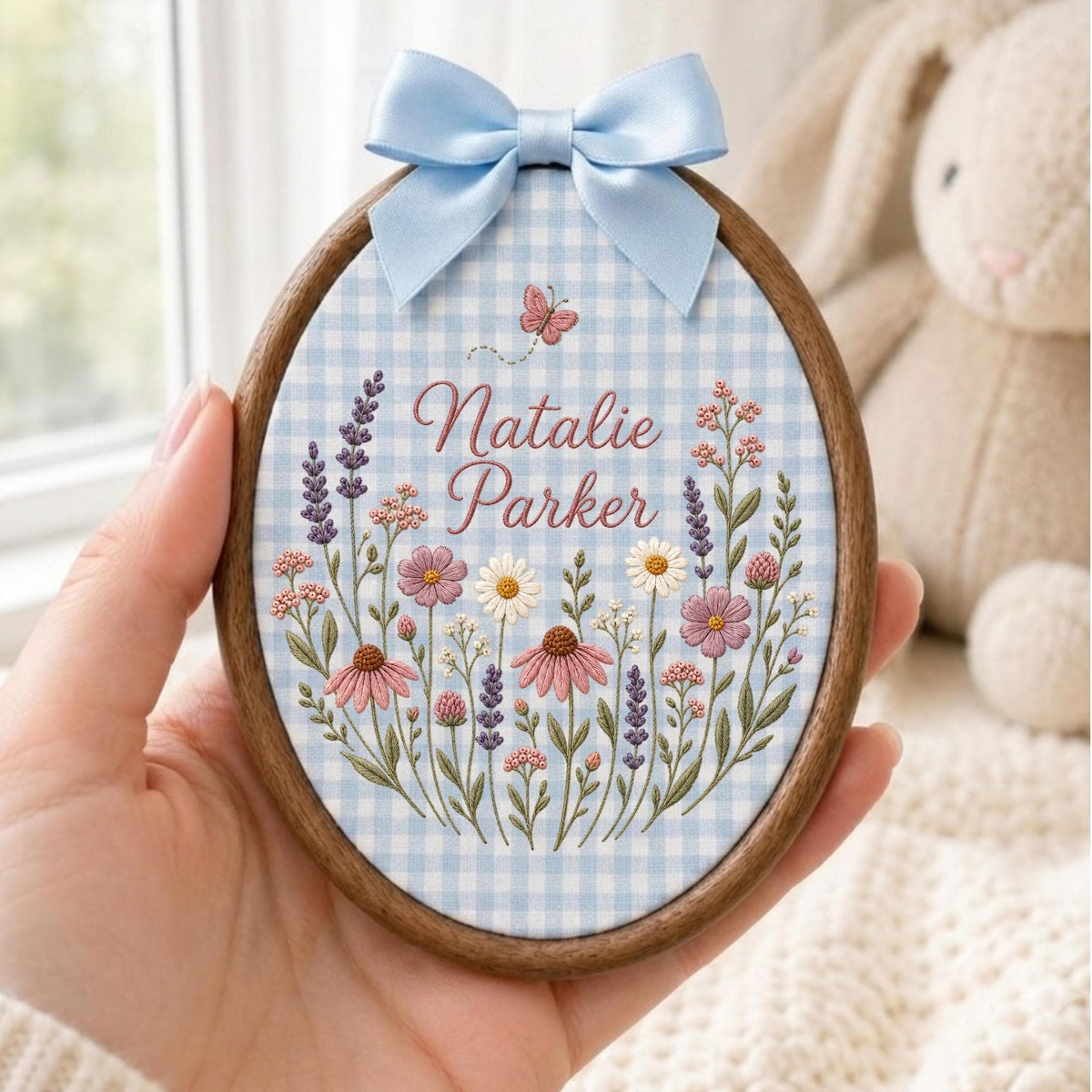 May include: An oval embroidery hoop featuring a light blue and white gingham fabric background. The center displays the name Natalie Parker in script text, surrounded by embroidered wildflowers in shades of pink, purple, and white, with a small butterfly above. A light blue satin ribbon bow is attached to the top of the dark wood frame.
