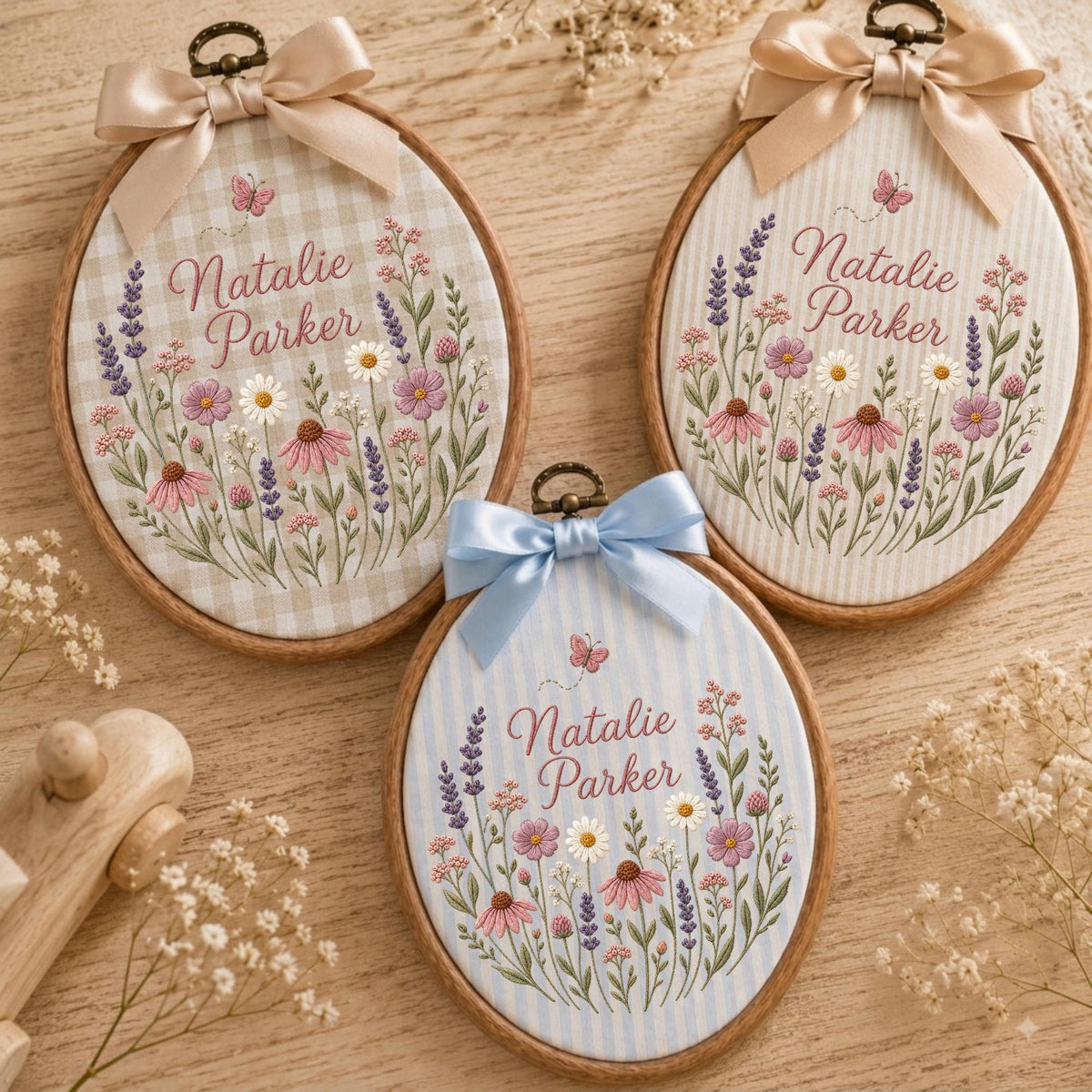May include: Three oval wooden embroidery hoops featuring fabric with floral patterns and the name Natalie Parker embroidered in script. Each hoop is topped with a satin ribbon bow in beige, light blue, or cream, displaying wildflowers.