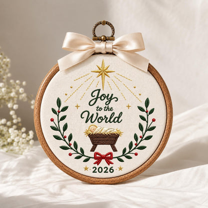 May include: A circular wooden embroidery hoop containing white fabric with a stitched design. The embroidery features a gold star, the text "Joy to the World", a manger with a baby, green holly branches with red berries, and the year "2026". A cream satin ribbon is tied at the top.