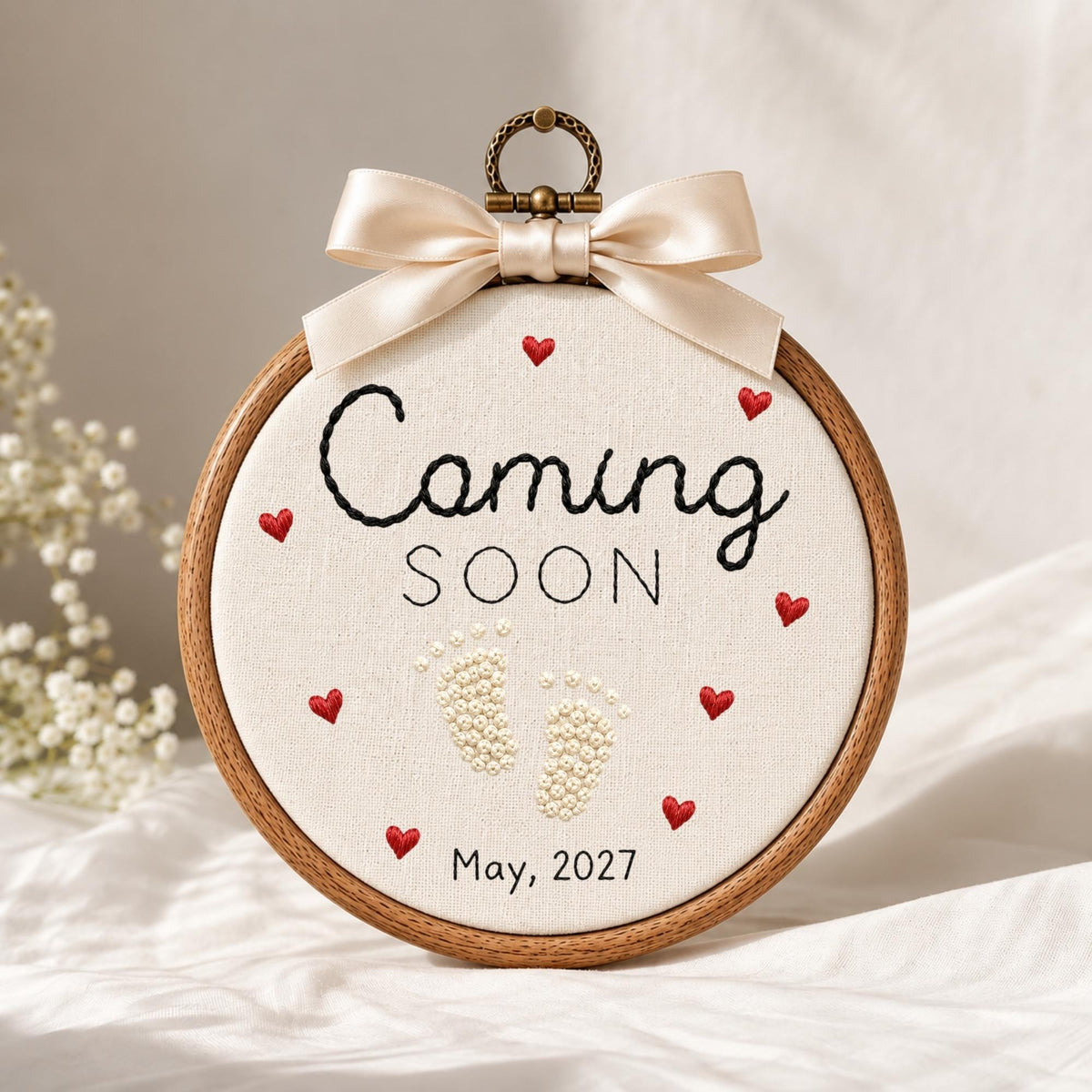 May include: A round wooden embroidery hoop containing white fabric with black embroidered text reading Coming SOON and May, 2027. The design features two small white embroidered baby footprints and several small red embroidered hearts. A beige satin bow is attached to the top metal hanging ring.
