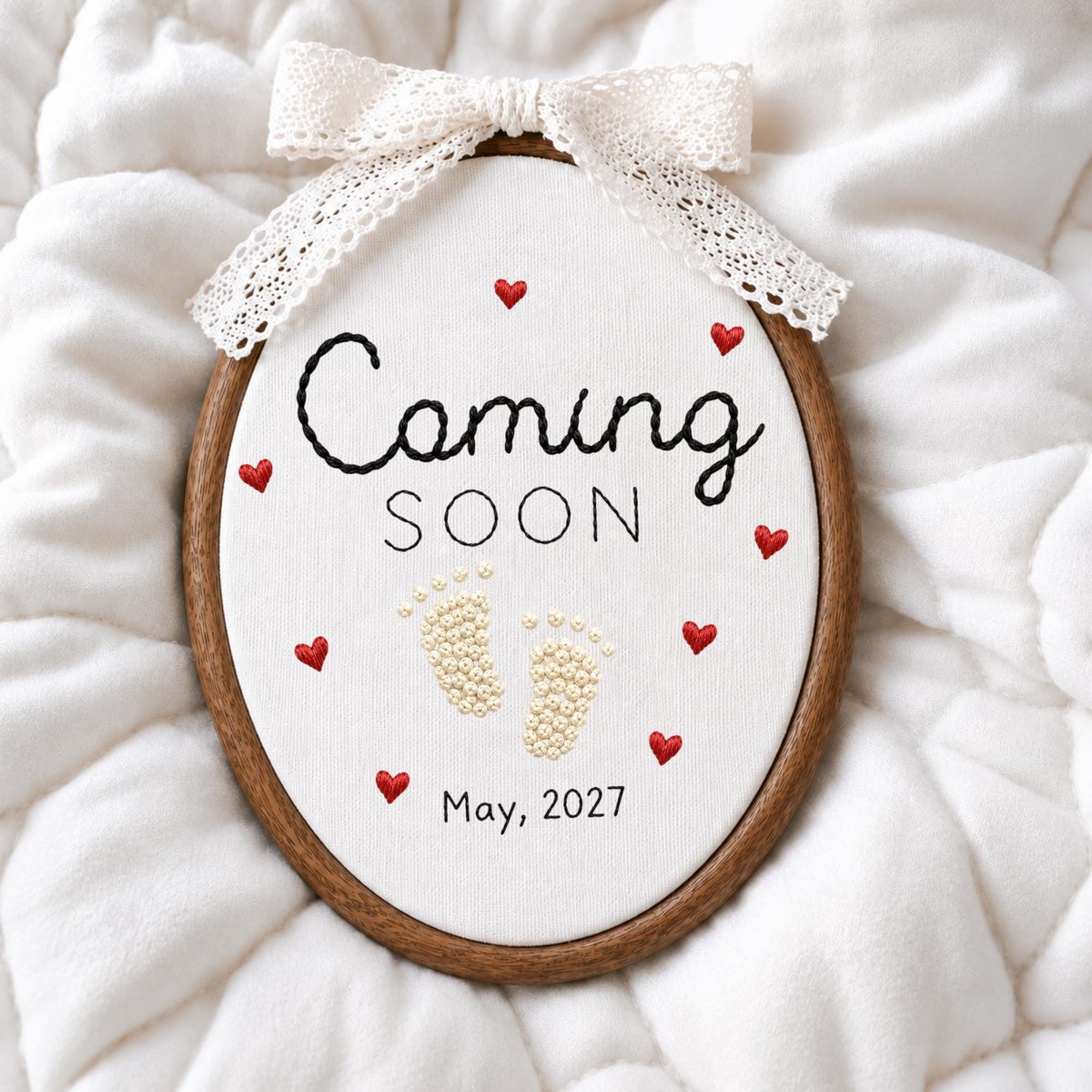 May include: An oval embroidery hoop containing white fabric with the black embroidered text Coming SOON and May, 2027. The design features small red embroidered hearts and cream-colored baby footprints. A white lace ribbon is tied at the top.