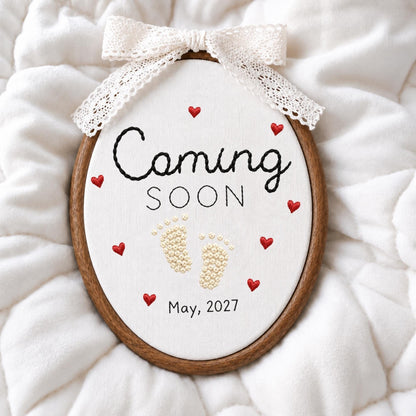 May include: An oval embroidery hoop containing white fabric with the black embroidered text Coming SOON and May, 2027. The design features small red embroidered hearts and cream-colored baby footprints. A white lace ribbon is tied at the top.