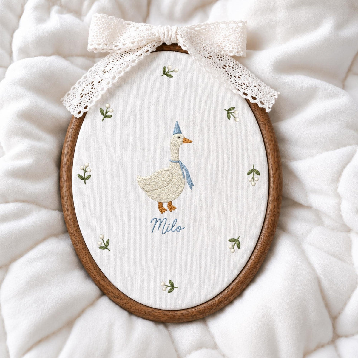 May include: An oval wooden embroidery hoop containing white fabric with a hand-stitched goose wearing a blue party hat and scarf. The name Milo is embroidered below the goose, surrounded by small floral sprigs. A cream lace bow tops the hoop.