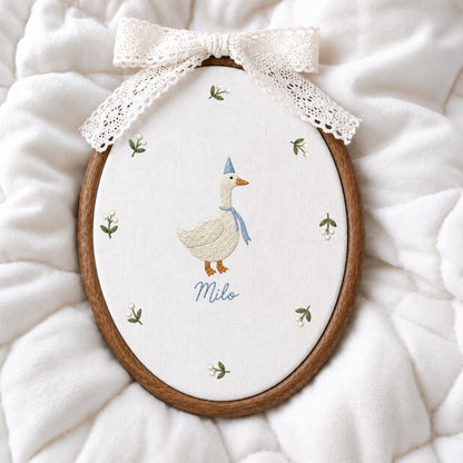 May include: An oval wooden embroidery hoop containing white fabric with a hand-stitched goose wearing a blue party hat and scarf. The name Milo is embroidered below the goose, surrounded by small floral sprigs. A cream lace bow tops the hoop.