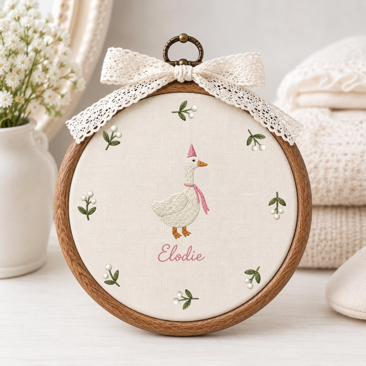 May include: A circular wooden embroidery hoop containing off-white fabric with a stitched white goose wearing a pink party hat and scarf. The name Elodie is embroidered in pink below the goose, surrounded by small green sprigs with white berries.