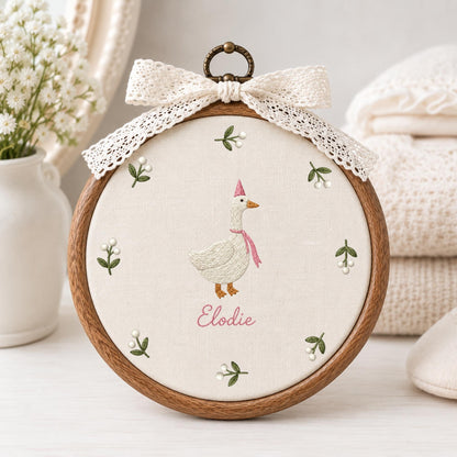 May include: A circular wooden embroidery hoop containing off-white fabric with a stitched white goose wearing a pink party hat and scarf. The name Elodie is embroidered in pink below the goose, surrounded by small green sprigs with white berries.