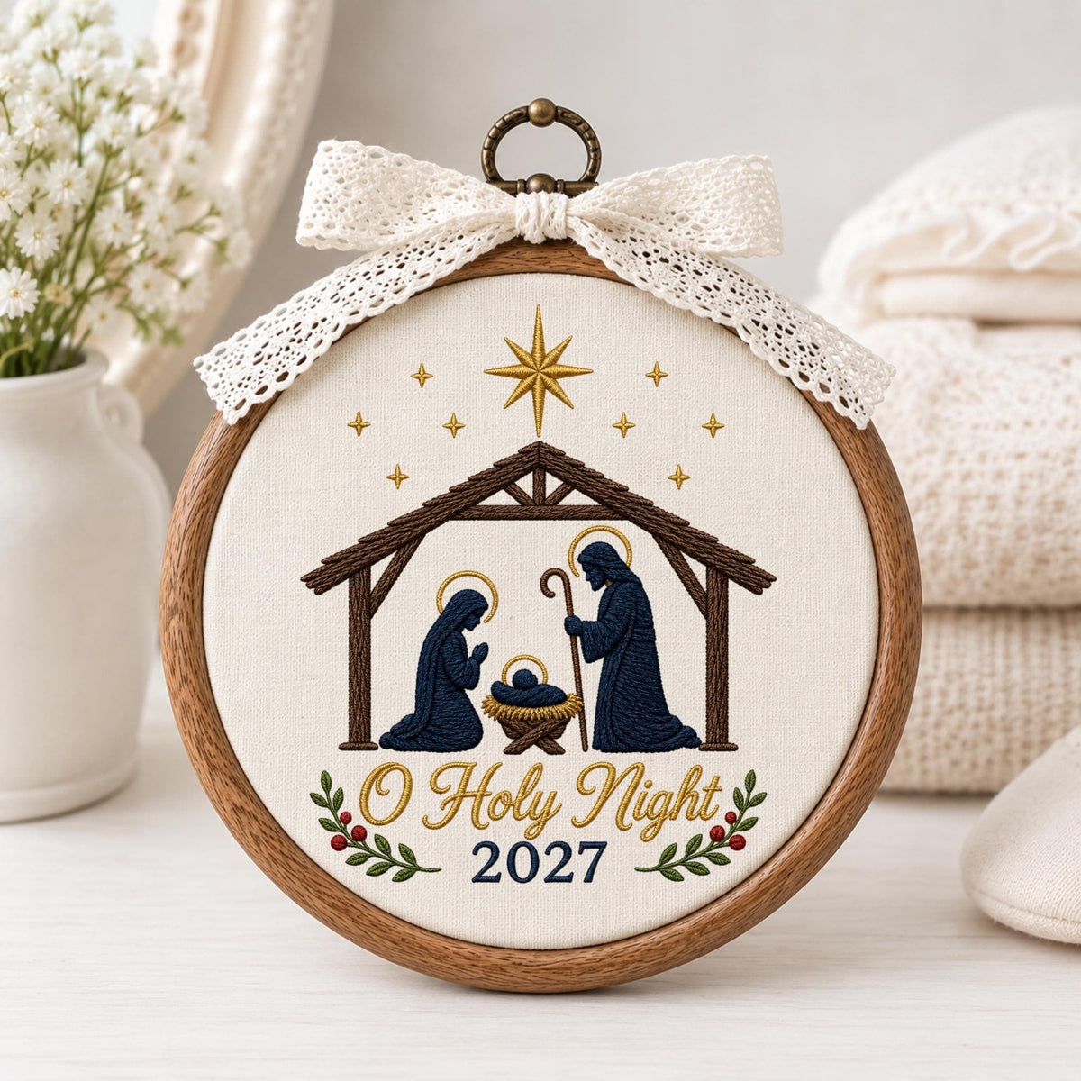 May include: A circular wooden embroidery hoop featuring a nativity scene with dark blue figures of Mary, Joseph, and the infant Jesus in a manger. Above is a gold star and smaller stars. Text reads O Holy Night and 2027, with a white lace bow.