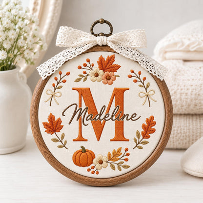 May include: A circular wooden embroidery hoop containing white fabric with a stitched design featuring a large orange letter M, the name Madeline in brown script, and an arrangement of embroidered autumn leaves, flowers, and a pumpkin.