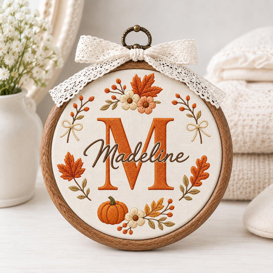 May include: A circular wooden embroidery hoop containing white fabric with a stitched design featuring a large orange letter M, the name Madeline in brown script, and an arrangement of embroidered autumn leaves, flowers, and a pumpkin.