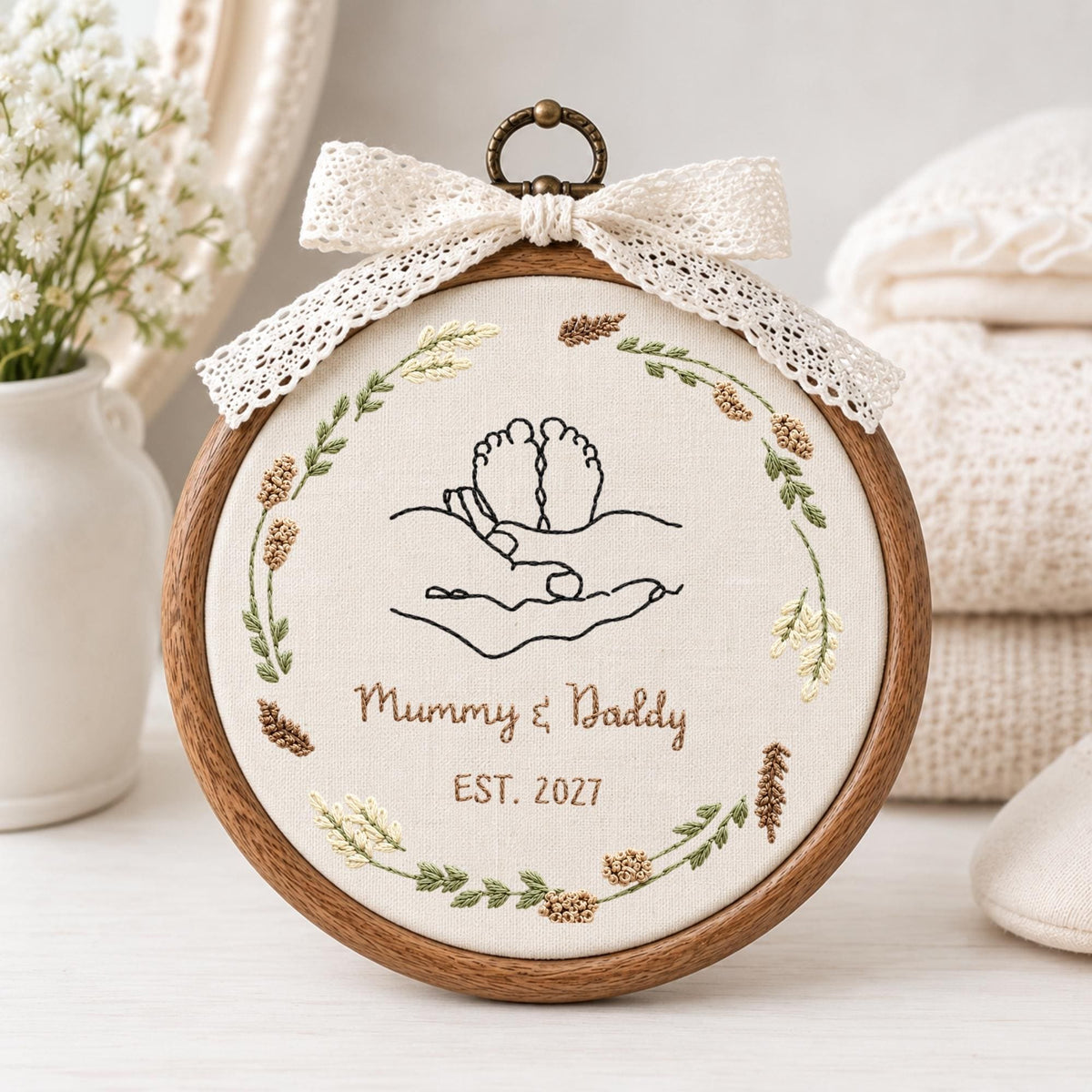 May include: A circular wooden embroidery hoop containing cream fabric with a black line art design of baby feet held in hands. The text reads Mummy &amp; Daddy EST. 2027. A floral wreath surrounds the design, and a white lace bow sits at the top.