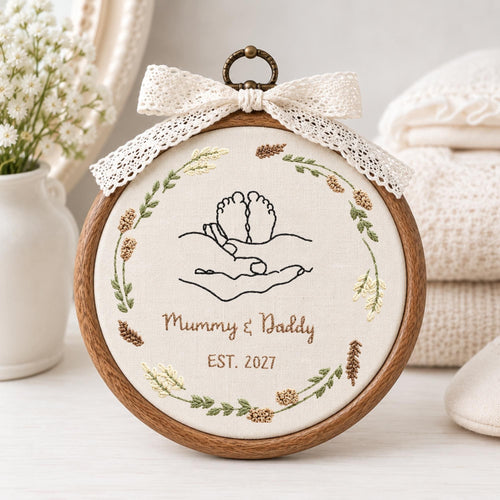 May include: A circular wooden embroidery hoop containing cream fabric with a black line art design of baby feet held in hands. The text reads Mummy &amp; Daddy EST. 2027. A floral wreath surrounds the design, and a white lace bow sits at the top.