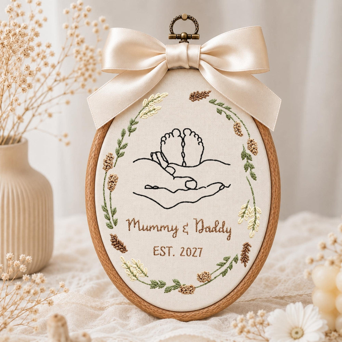 May include: An oval embroidery hoop featuring a cream fabric background with black line art of hands cradling baby feet. The design is framed by a botanical wreath in green and brown tones. Below, the text reads Mummy &amp; Daddy EST. 2027. A large cream satin bow is attached to the top.