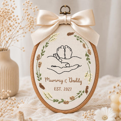 May include: An oval embroidery hoop featuring a cream fabric background with black line art of hands cradling baby feet. The design is framed by a botanical wreath in green and brown tones. Below, the text reads Mummy &amp; Daddy EST. 2027. A large cream satin bow is attached to the top.