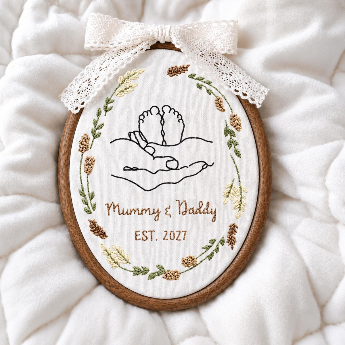 May include: An oval embroidery hoop containing white fabric with black line art of baby feet held in hands. The design is surrounded by a wreath of embroidered wheat and leaves. Below, brown text reads Mummy &amp; Daddy EST. 2027. A white lace bow is attached to the top.