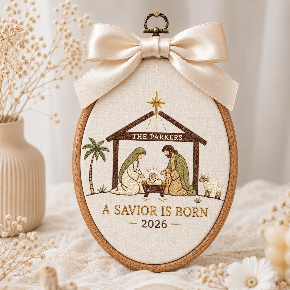 May include: An oval wooden embroidery hoop featuring a nativity scene with embroidered figures of Mary, Joseph, and the infant Jesus. The text reads THE PARKERS, A SAVIOR IS BORN, and 2026. A large cream-colored satin ribbon bow is attached to the top.