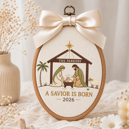 May include: An oval wooden embroidery hoop featuring a nativity scene with embroidered figures of Mary, Joseph, and the infant Jesus. The text reads THE PARKERS, A SAVIOR IS BORN, and 2026. A large cream-colored satin ribbon bow is attached to the top.