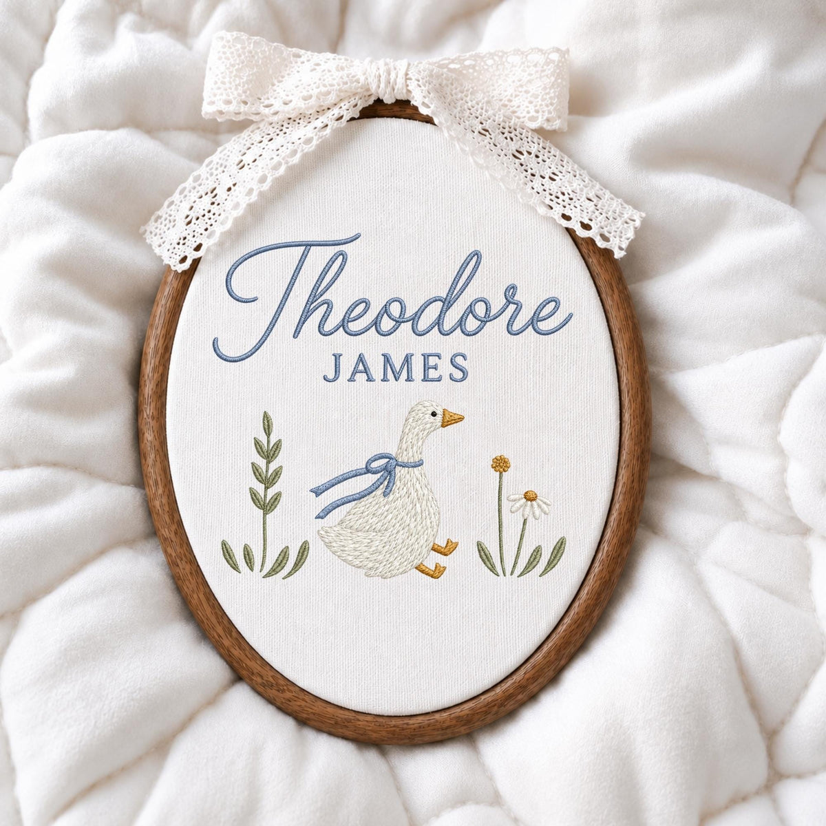 May include: An oval embroidery hoop featuring a white fabric background with the embroidered blue text Theodore JAMES. Below the text is a white goose with a blue ribbon around its neck, flanked by green leafy stems and a small white flower.