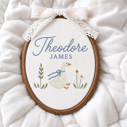 May include: An oval embroidery hoop featuring a white fabric background with the embroidered blue text Theodore JAMES. Below the text is a white goose with a blue ribbon around its neck, flanked by green leafy stems and a small white flower.