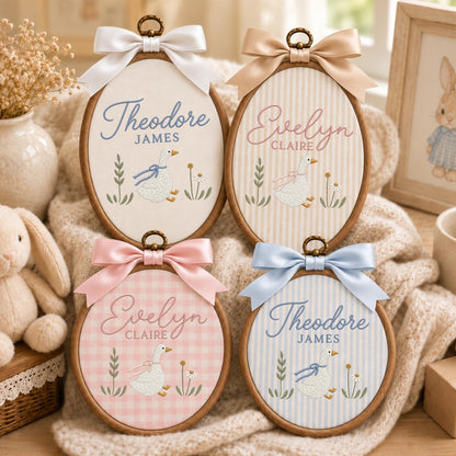May include: Four oval wooden embroidery hoops featuring fabric inserts with embroidered geese, floral accents, and names including Theodore James and Evelyn Claire. Each hoop is topped with a decorative satin ribbon bow in white, tan, pink, or blue.