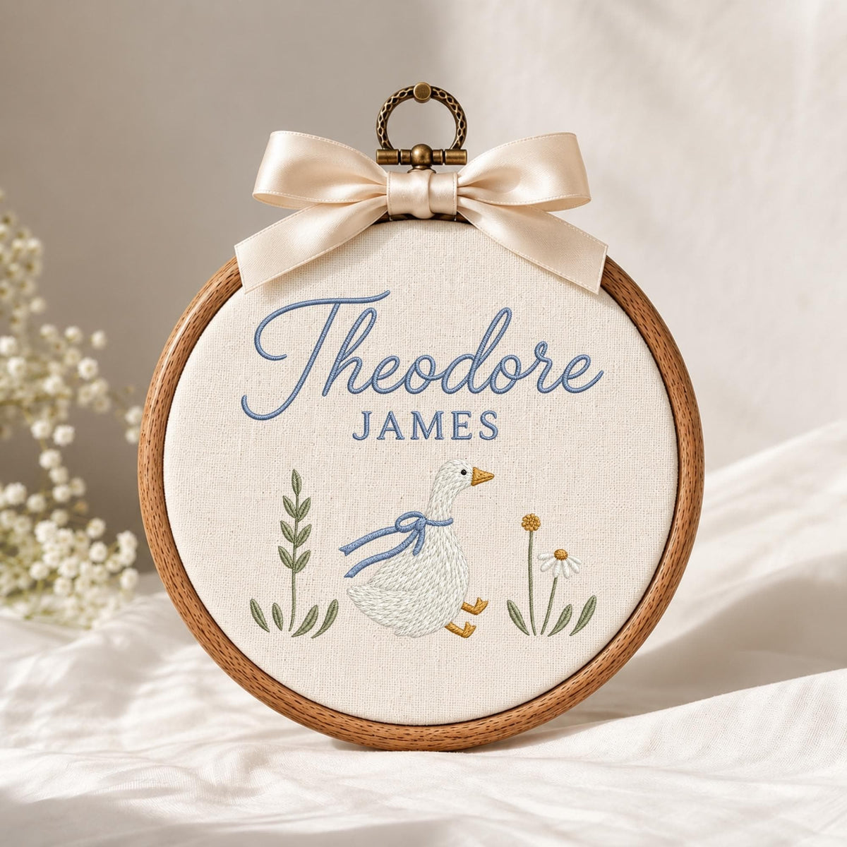 May include: A circular wooden embroidery hoop containing cream-colored fabric with the embroidered name Theodore JAMES in blue. Below the text is a white goose with a blue ribbon, green leafy stems, and a small yellow and white flower.