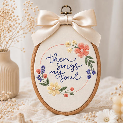 May include: An oval wooden embroidery hoop containing cream fabric with the embroidered dark blue text then sings my soul. The design features a floral wreath of pink, yellow, and blue flowers with green leaves, topped with a large cream satin bow.