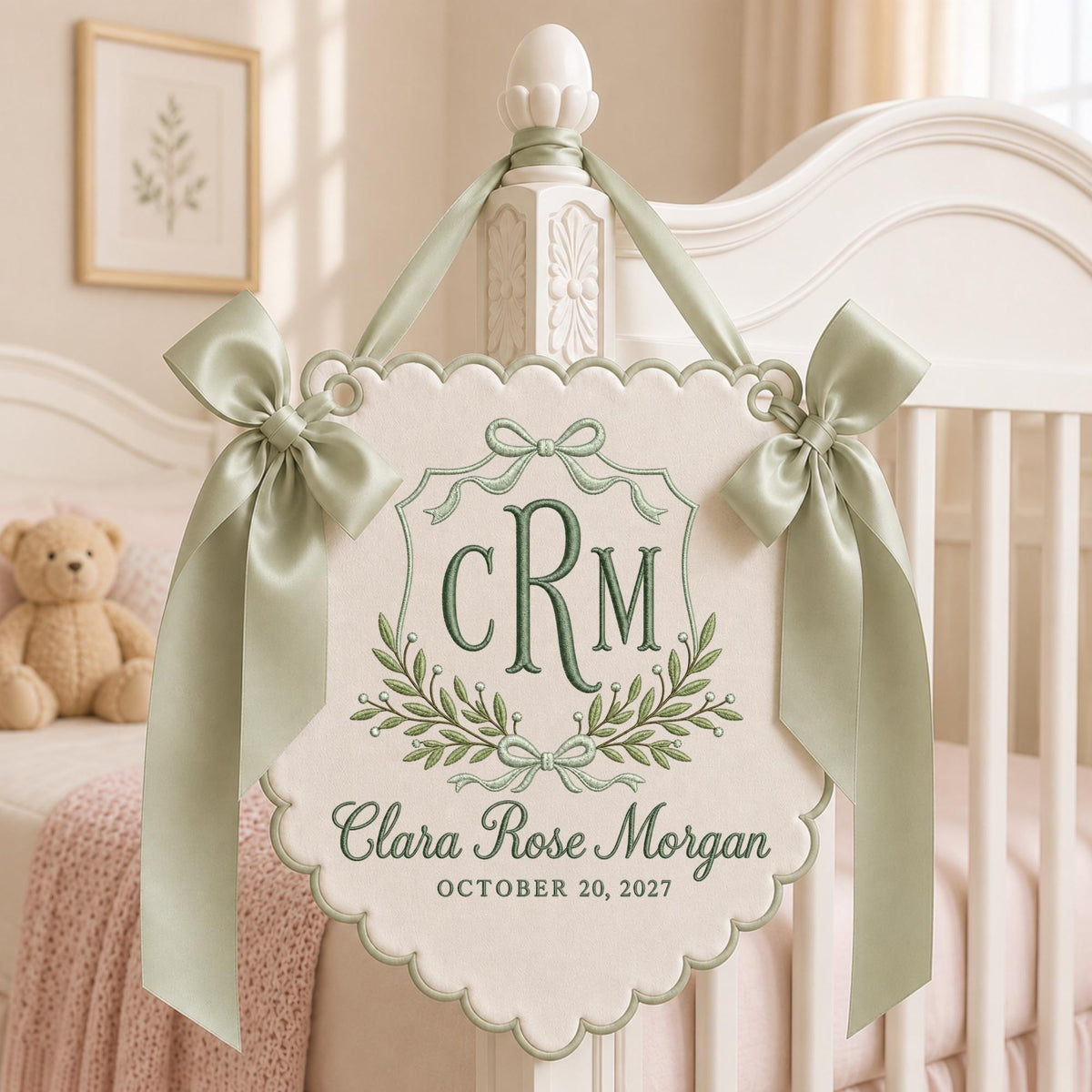 May include: A cream-colored fabric wall hanging with scalloped edges, featuring sage green embroidery of the monogram CRM, the name Clara Rose Morgan, and the date October 20, 2027. It is adorned with two sage green satin ribbons tied in bows.