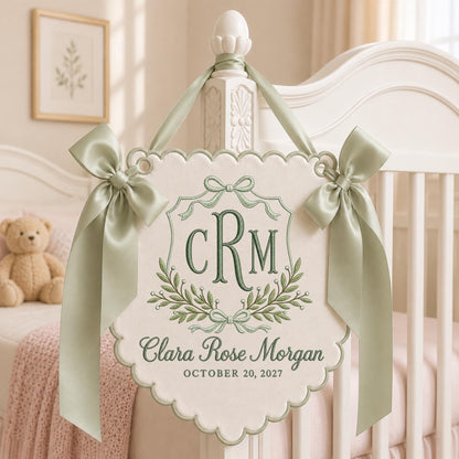 May include: A cream-colored fabric wall hanging with scalloped edges, featuring sage green embroidery of the monogram CRM, the name Clara Rose Morgan, and the date October 20, 2027. It is adorned with two sage green satin ribbons tied in bows.