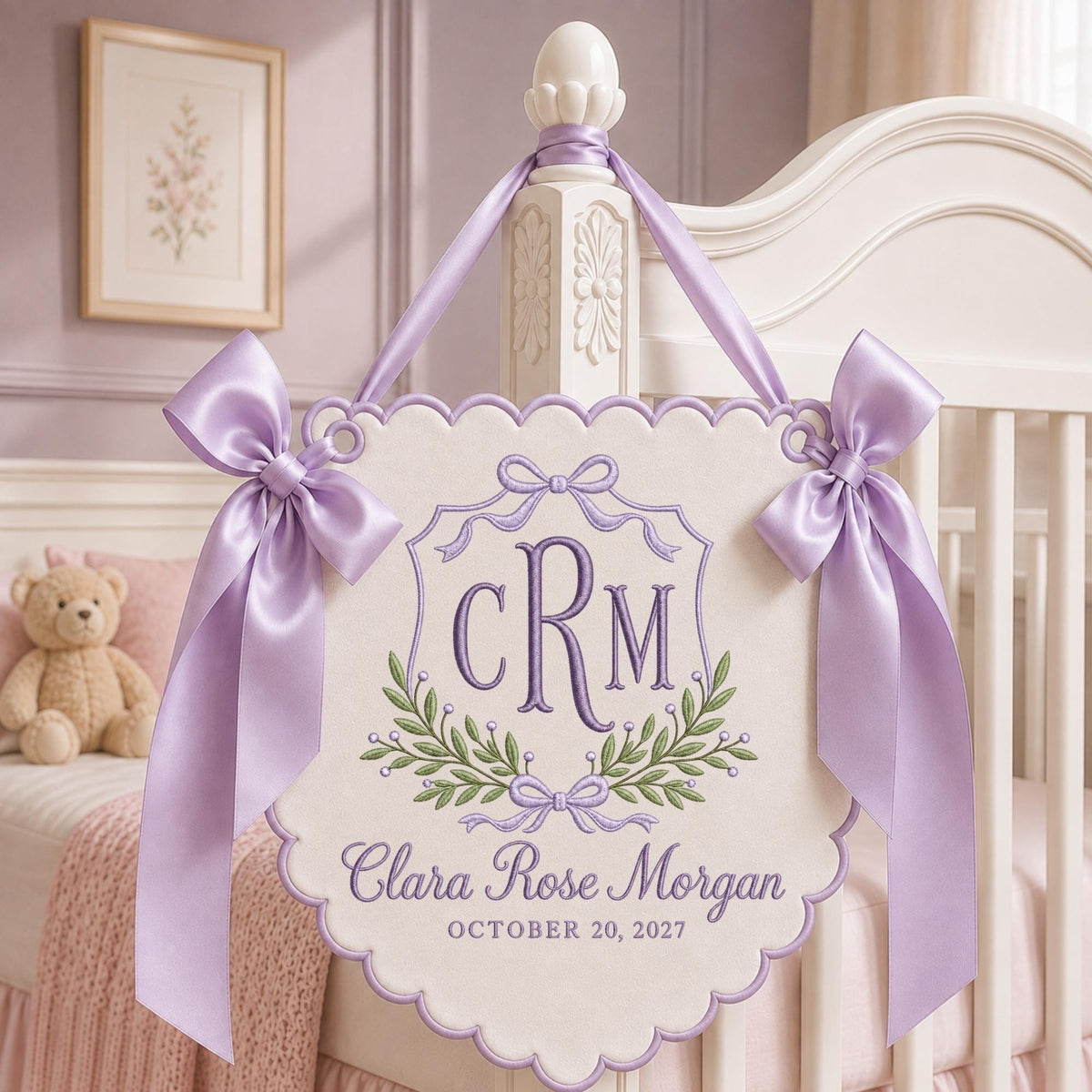 May include: A cream-colored scalloped wall hanging featuring purple embroidery of the monogram CRM, a laurel wreath, and the name Clara Rose Morgan with the date October 20, 2027. The sign is accented with two large lavender satin bows and ribbons.
