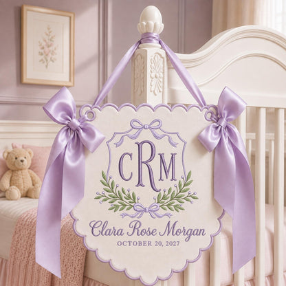 May include: A cream-colored scalloped wall hanging featuring purple embroidery of the monogram CRM, a laurel wreath, and the name Clara Rose Morgan with the date October 20, 2027. The sign is accented with two large lavender satin bows and ribbons.
