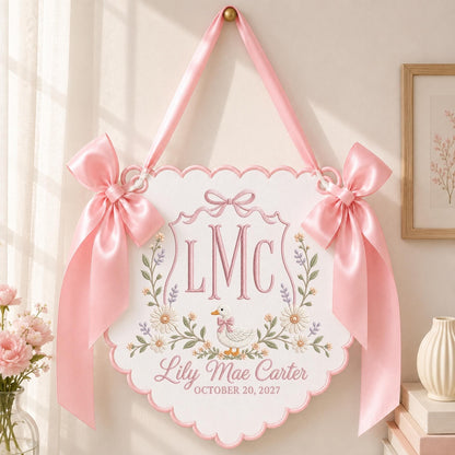 May include: A white scalloped wall hanging featuring pink embroidered text LMC, the name Lily Mae Carter, and the date October 20, 2027. The design includes a central duck illustration, floral accents, and two large pink satin ribbon bows.