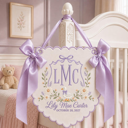 May include: A scalloped, off-white fabric wall hanging featuring purple embroidery of the monogram LMC, a duck, and floral accents. The text reads Lily Mae Carter and October 20, 2027. It is adorned with two large, light purple satin ribbon bows.