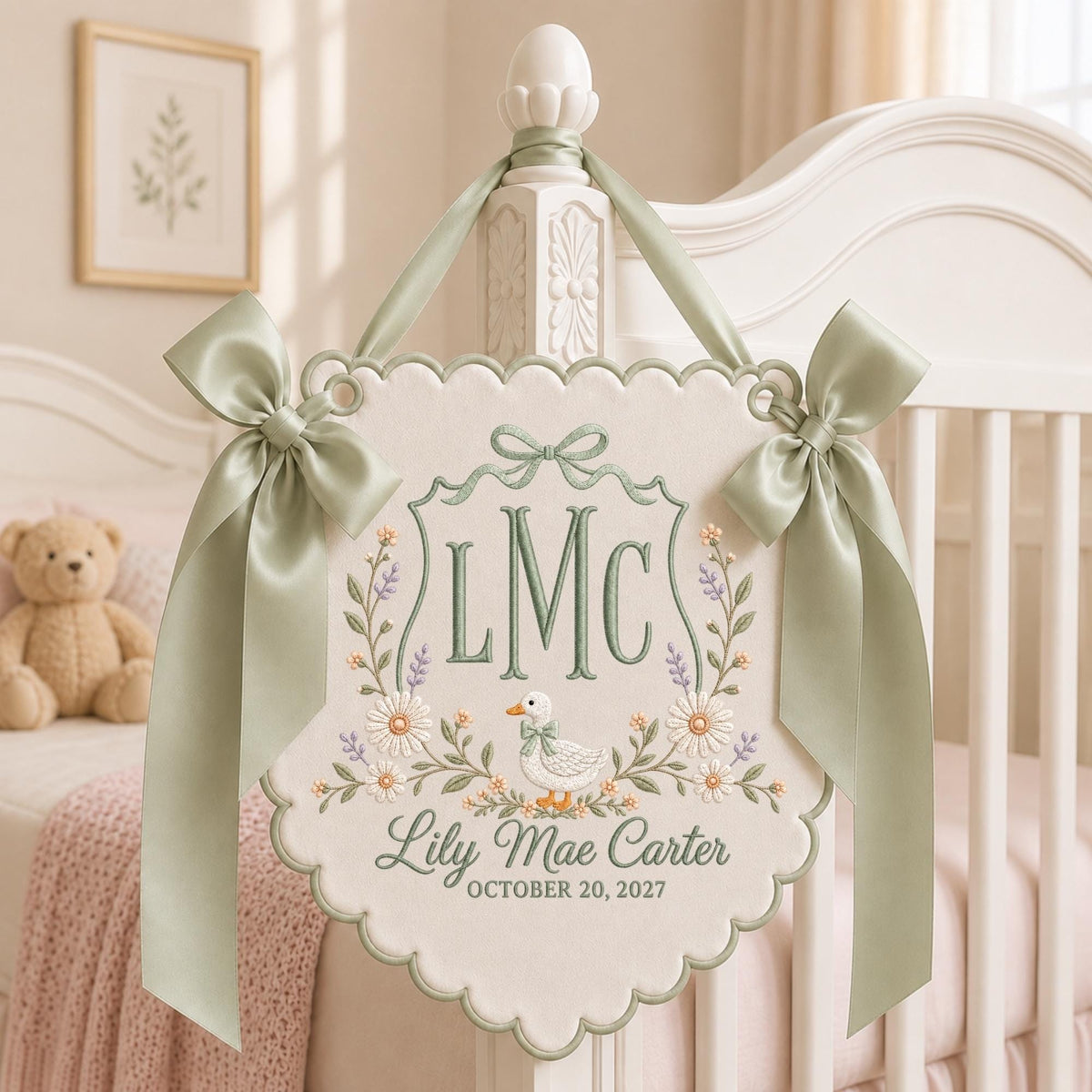 May include: A cream-colored fabric wall hanging with scalloped edges, featuring sage green embroidery of the initials LMC, a duck, and floral accents. It includes the name Lily Mae Carter and the date OCTOBER 20, 2027, with sage green ribbon bows.
